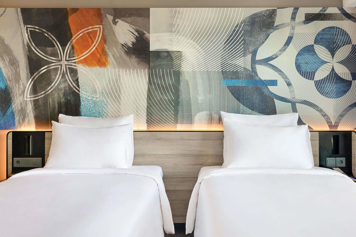 Ein Hotelzimmer mit zwei Betten, einem modernen, abstrakten Wandbild und einem hölzernen Kopfteil.