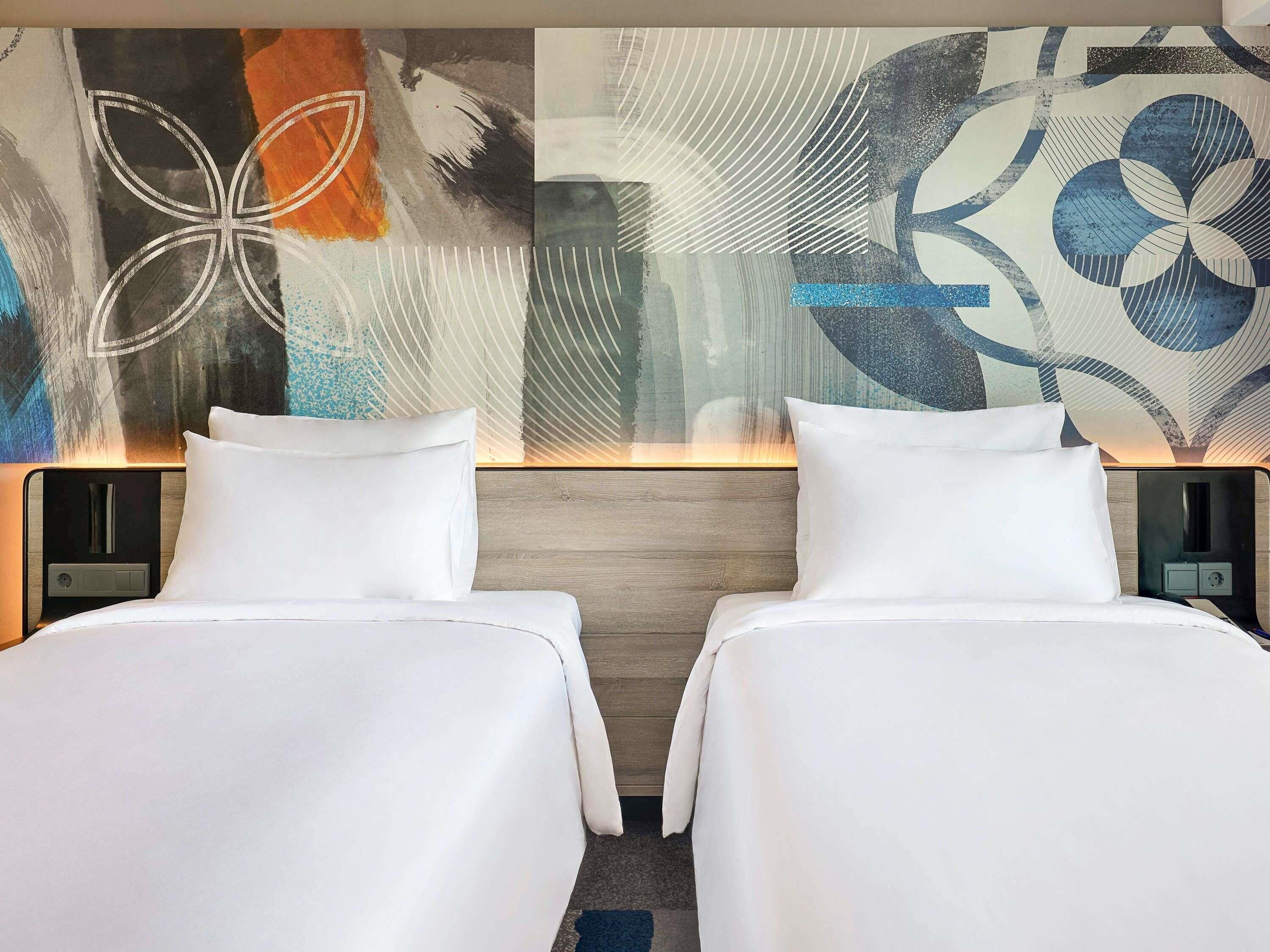 Ein Hotelzimmer mit zwei Betten, einem modernen, abstrakten Wandbild und einem hölzernen Kopfteil.
