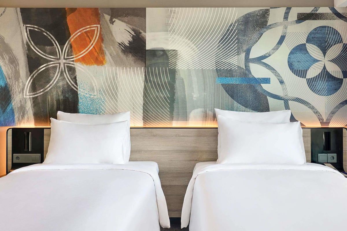 A hotel room with two beds, a modern abstract wall art, and a wooden headboard.