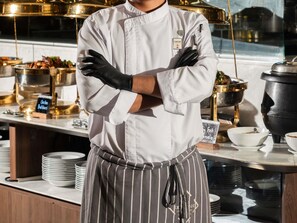 Een kok in een wit uniform met een gestreept schort, gekruiste armen in een keuken.