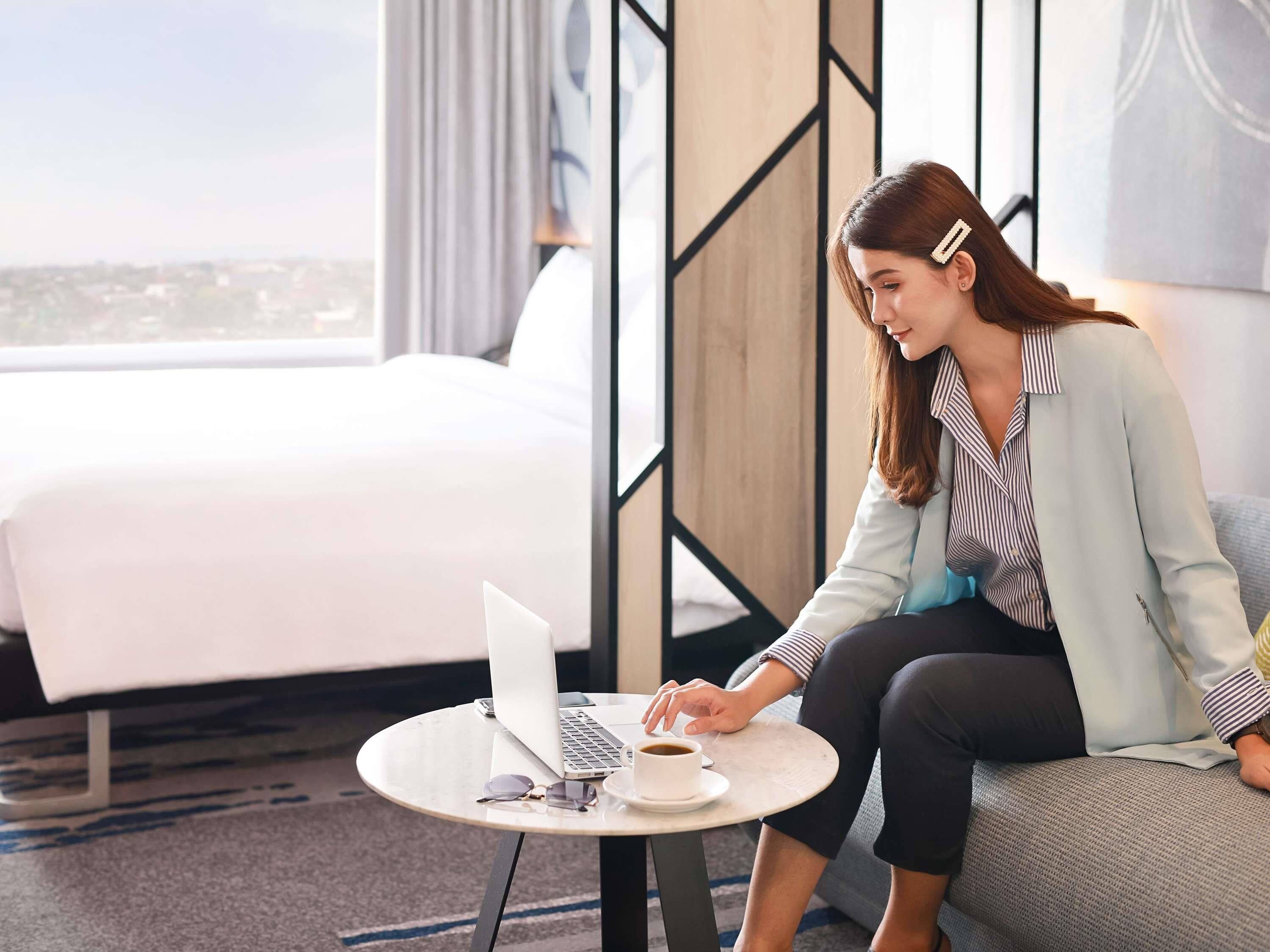 A person sitting on a couch with a laptop and a cup of coffee, in a room with a bed and a view of the city.