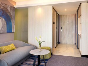 A modern hotel room with a grey sofa, a round table with a vase of flowers, and a hallway with wooden doors.