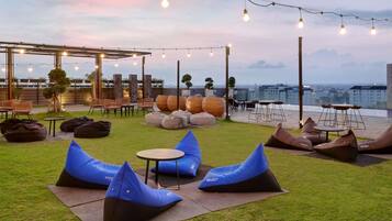An outdoor seating area with bean bags, tables, and chairs, under string lights, with a city view in the background.