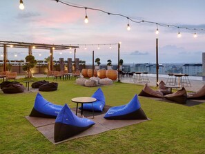 An outdoor seating area with bean bags, tables, and chairs, under string lights, with a city view in the background. - Novotel Suites Yogyakarta Malioboro (Yogyakarta)