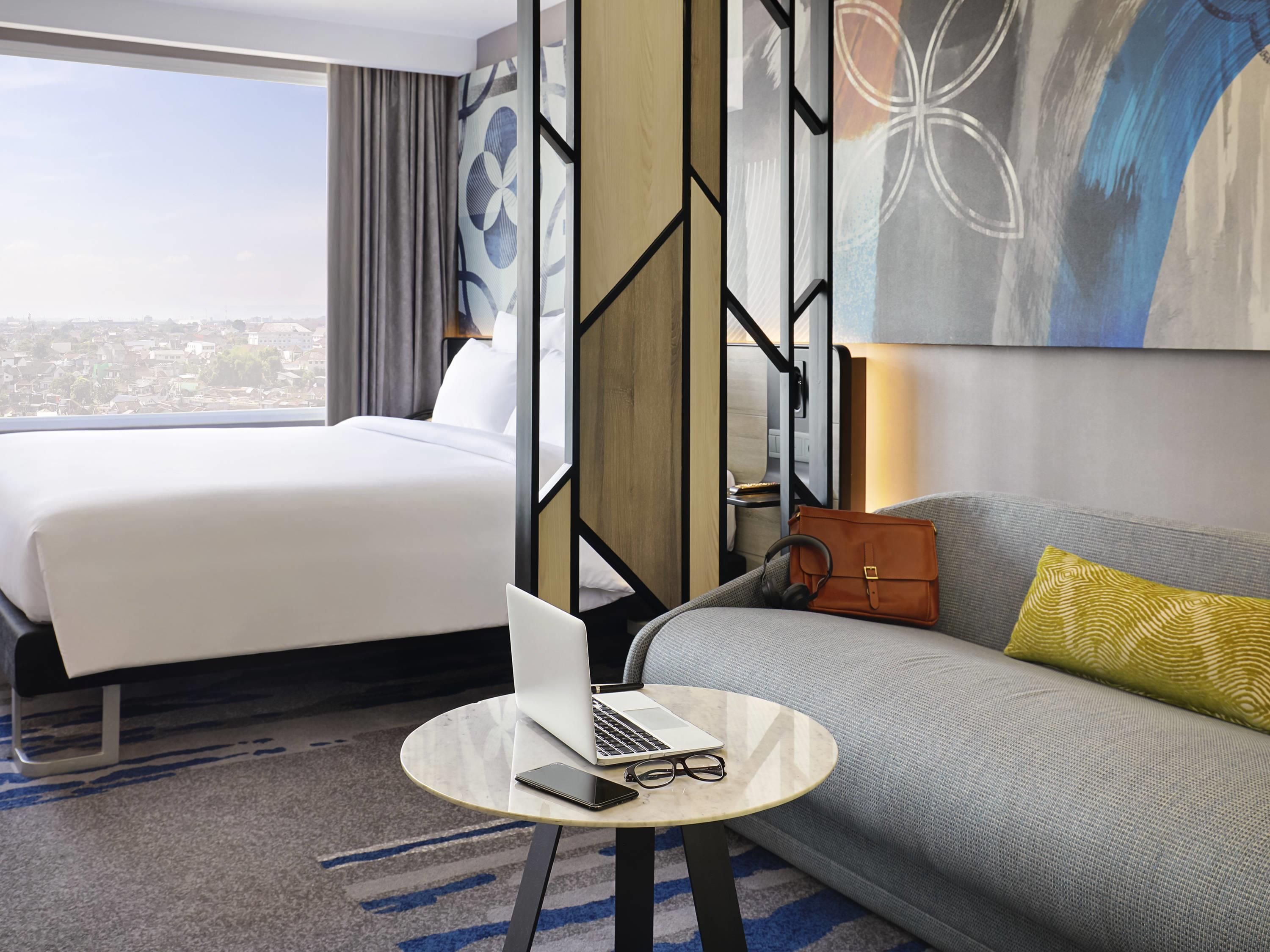Habitación de hotel con cama, sofá, una mesa redonda con una computadora portátil y vasos, y vista a la ciudad.