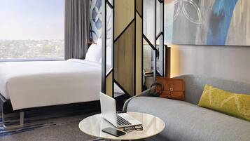Habitación de hotel con cama, sofá, una mesa redonda con una computadora portátil y vasos, y vista a la ciudad.