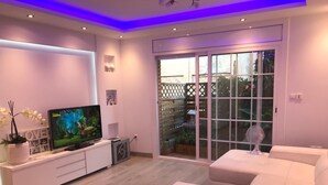 A modern living room with a flat-screen TV, white furniture, and a sliding glass door leading to a garden. - Kamelly Guest House (Badalona)
