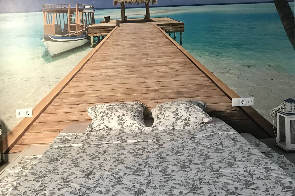 Eine Holzsteige führt zu einer Reetdachkate und einem Boot, im Hintergrund ein tropischer Strand und Palmen.