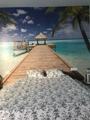 A wooden pier leading to a thatched hut and a boat, with a tropical beach and palm trees in the background. - Kamelly Guest House (Badalona)