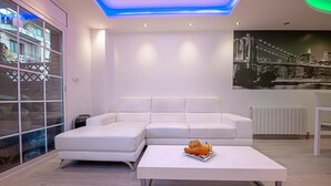A modern living room with a white sofa, a white coffee table with a fruit arrangement, and a cityscape mural on the wall. - Kamelly Guest House (Badalona)