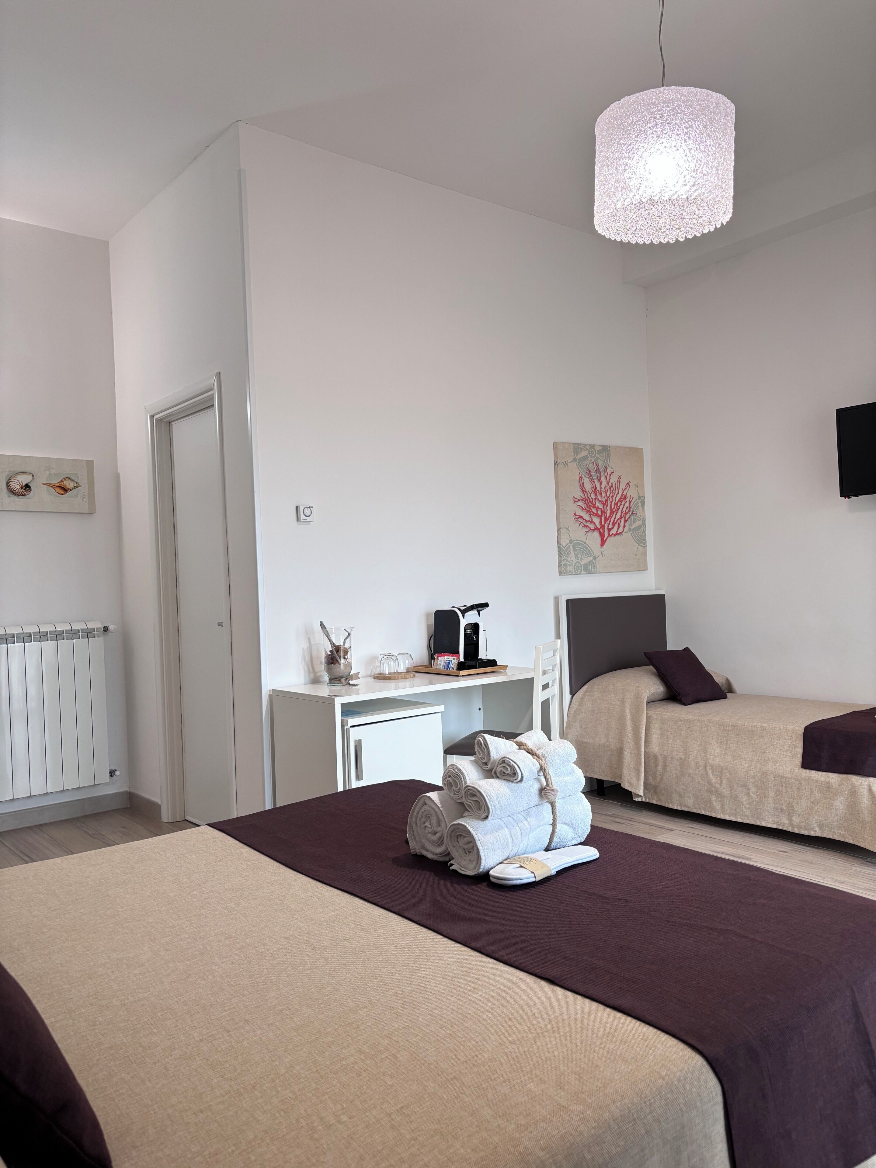 A hotel room with two beds, a desk with a coffee maker, a radiator, and a wall-mounted TV.