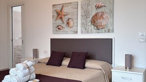 A neatly made bed with a dark headboard, a bedside table with a lamp, and two wall-mounted artworks depicting seashells and a starfish. - Faro Bianco Gallipoli (Gallipoli)