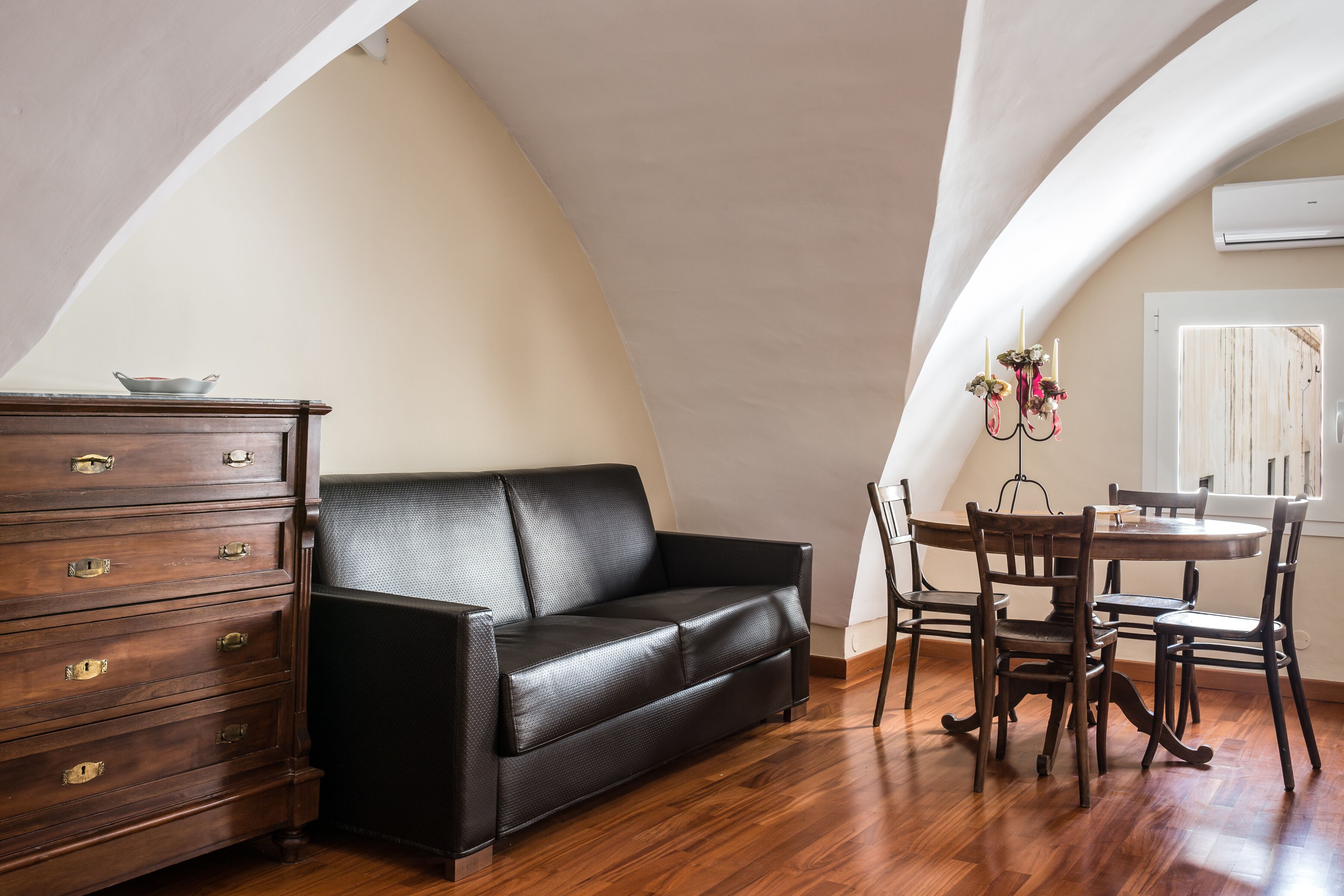 Une pièce comprenant un canapé en cuir noir, une commode en bois, une table à manger avec des chaises et un climatiseur blanc.