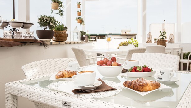 A breakfast setting with wicker chairs, a glass table, white coffee cups, croissants, a bowl of fruit, and hanging planters. - Faro Bianco Gallipoli (Gallipoli)