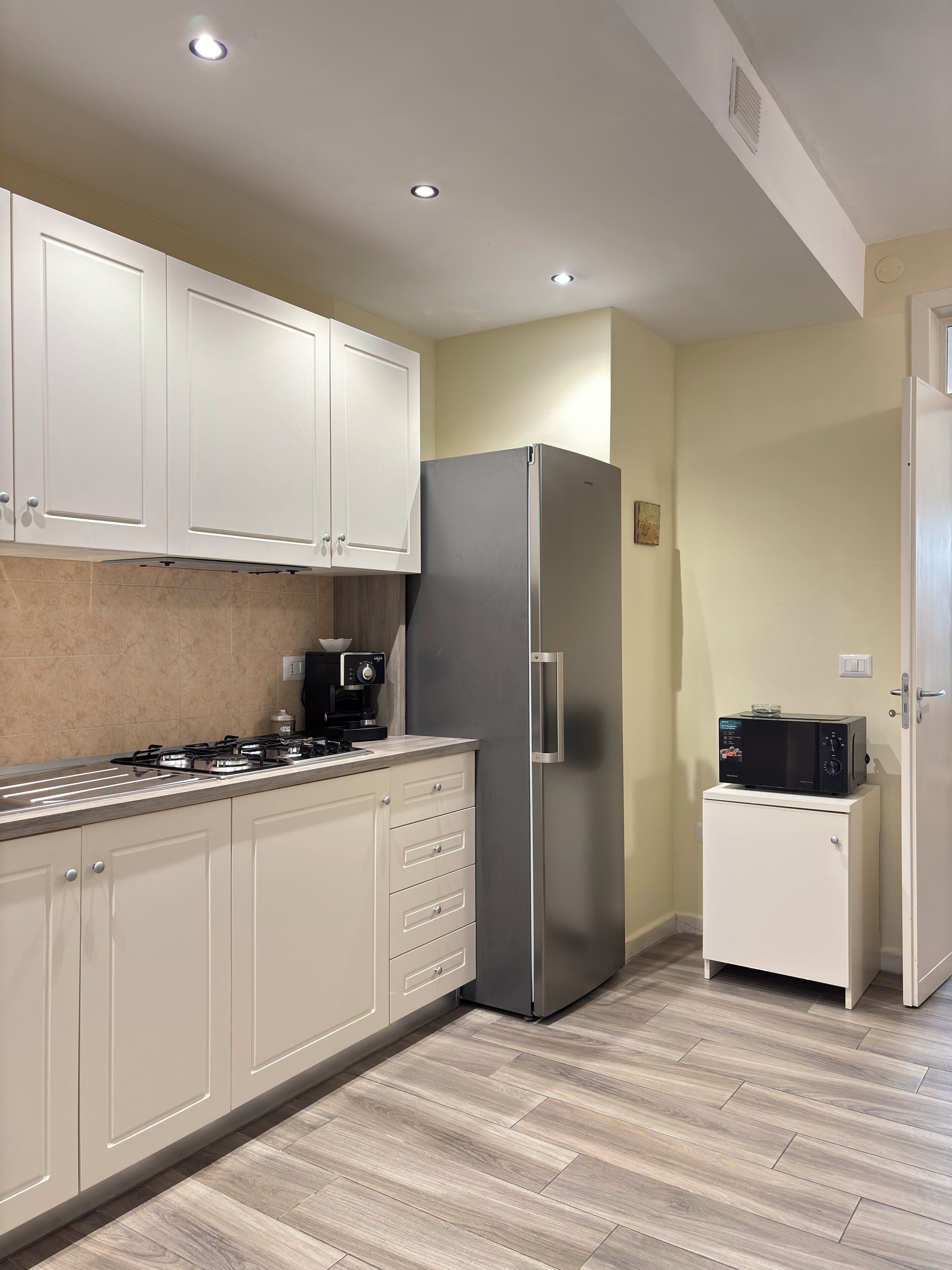 Cocina compacta con armarios blancos, un refrigerador de acero inoxidable, un microondas sobre un mueble pequeño y una estufa a gas.