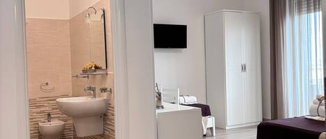 A modern hotel room with a wooden floor, a bed, a white wardrobe, a television, and a bathroom with a bidet and sink.