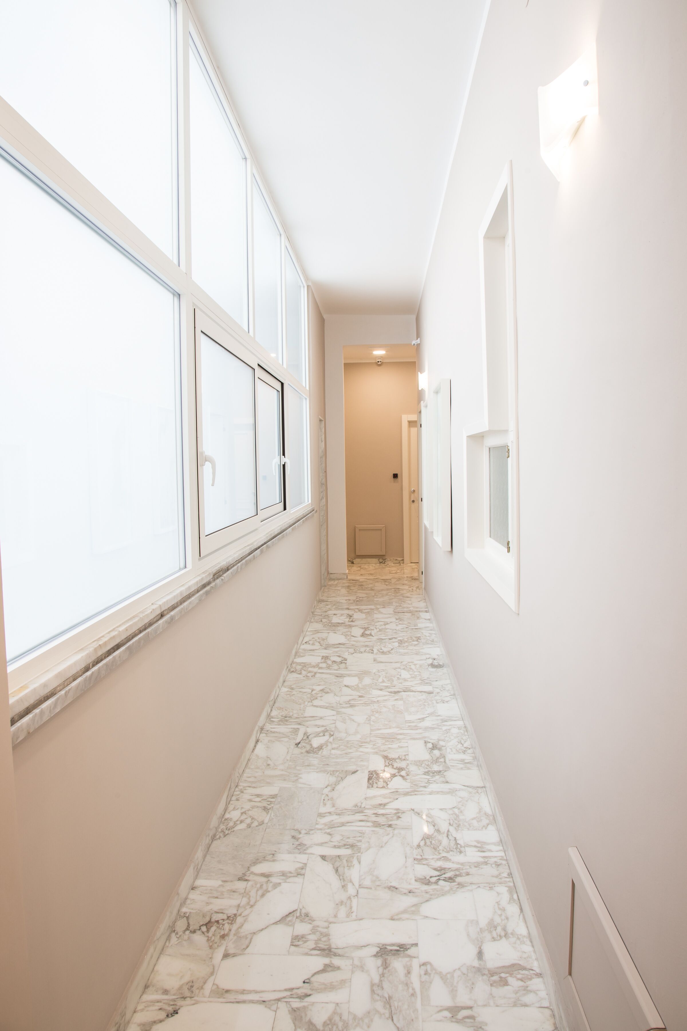 A narrow hallway with marble flooring, large windows, and framed pictures on the walls.