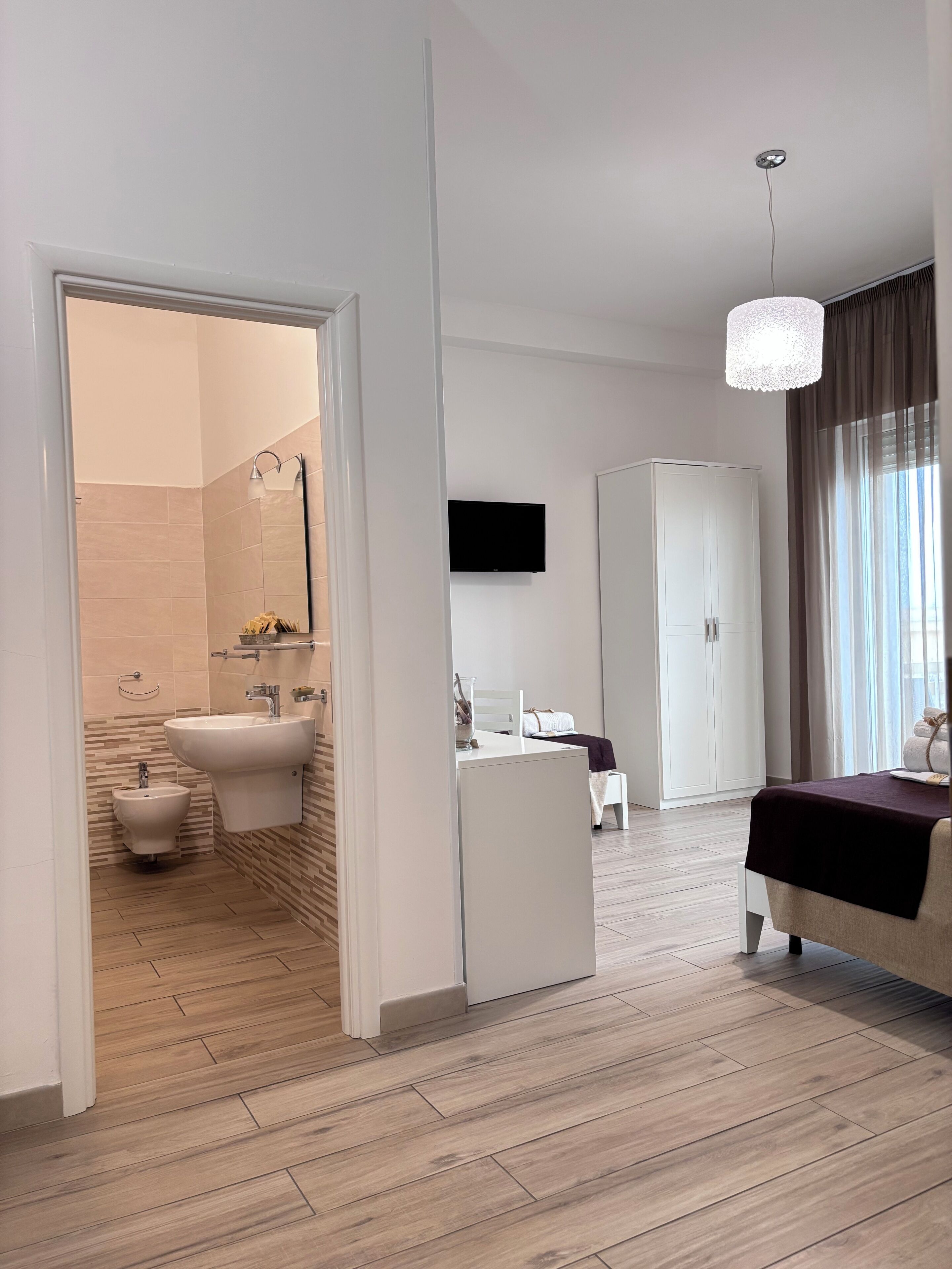 A modern hotel room with a wooden floor, a bed, a TV, a wardrobe, and a bathroom with a bidet and sink.