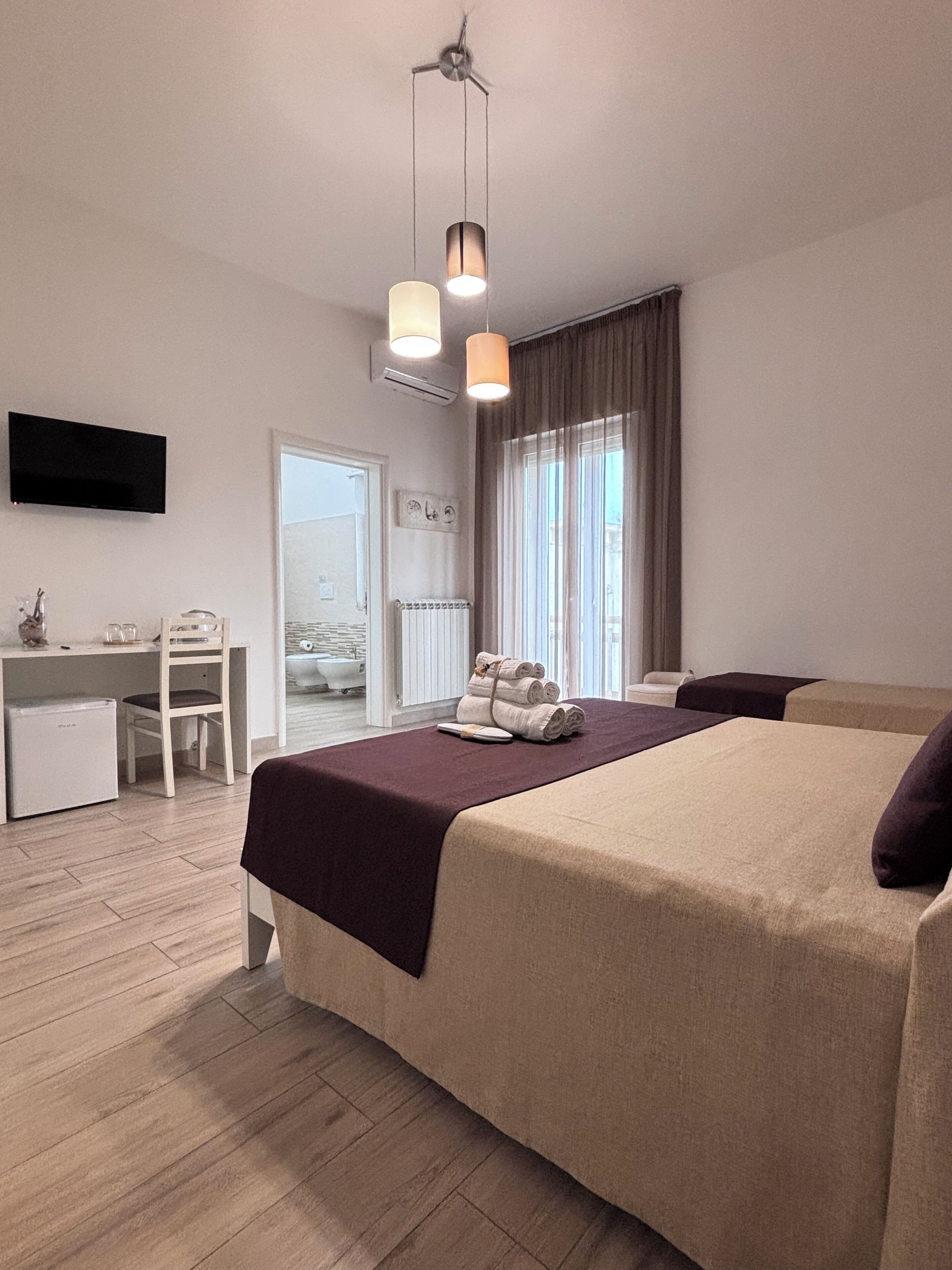 Habitación de hotel con cama, escritorio, silla, televisor y ventana con cortinas.