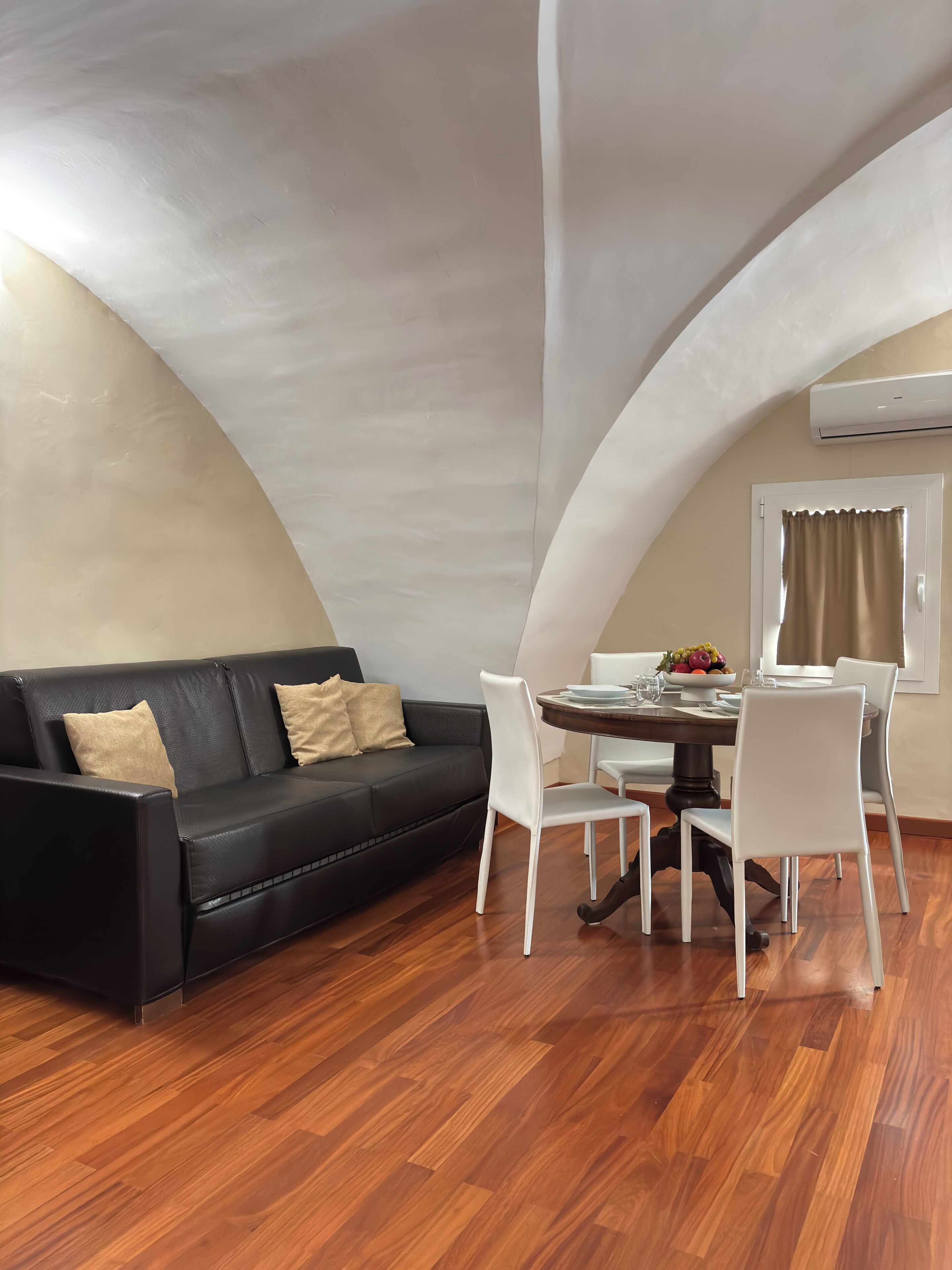 Ein modernes Wohnzimmer mit einem schwarzen Ledersofa, weißen Stühlen, einem Esstisch mit Obstschale und einem Fenster mit Vorhängen.