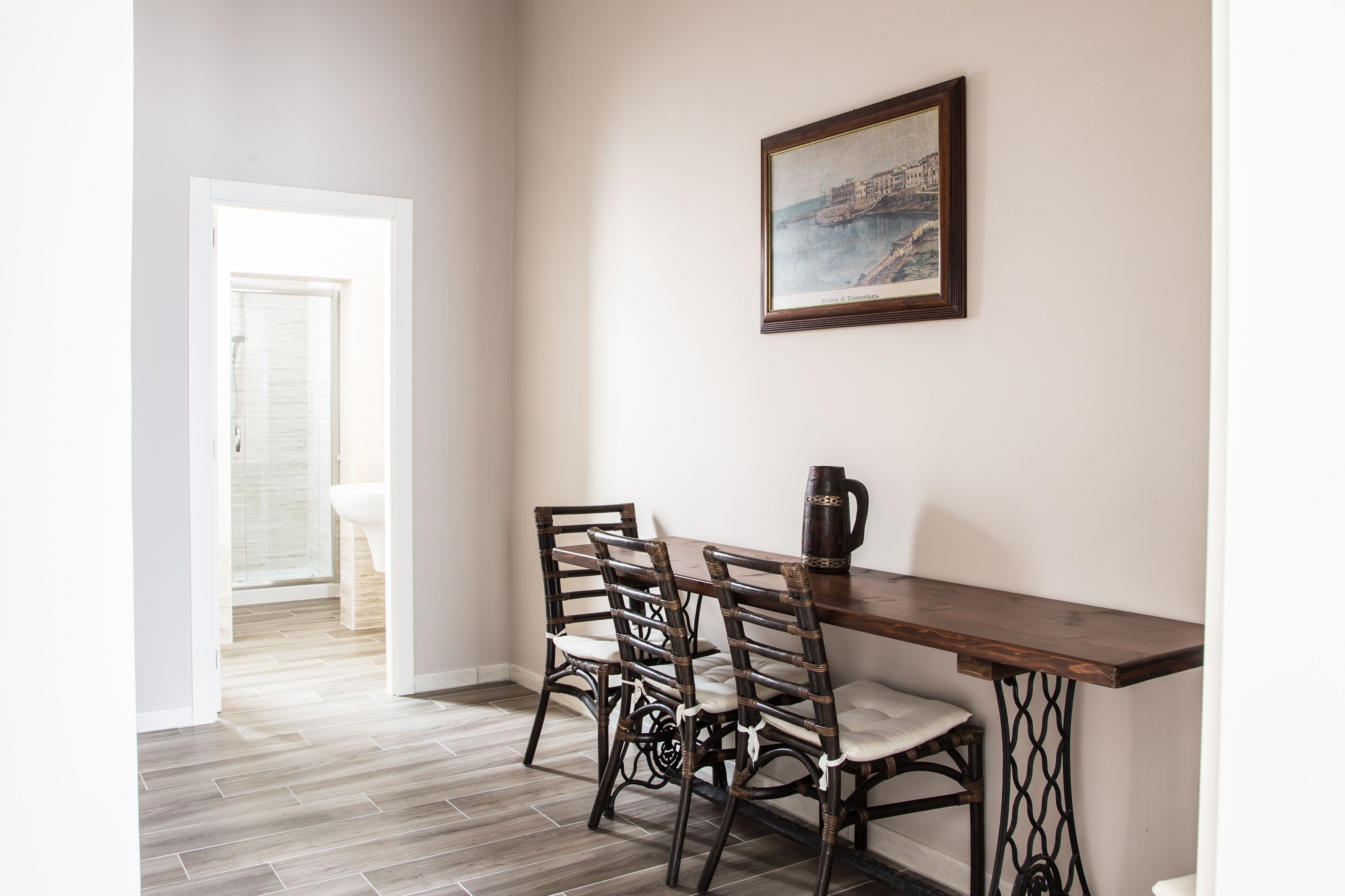 Un coin repas avec une table en bois et quatre chaises, un tableau encadré au mur et une vue sur une salle de bain par une porte ouverte.