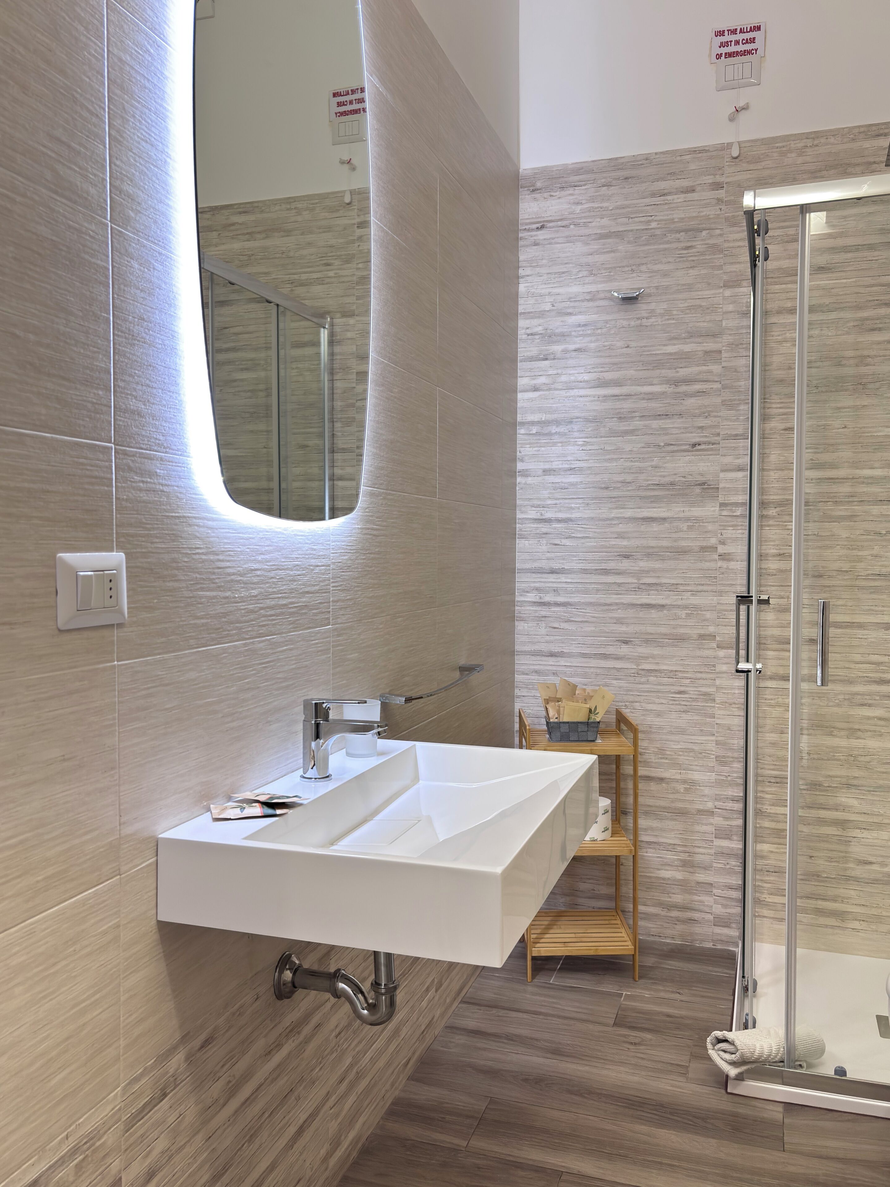 A modern bathroom with a large sink, mirror, shower, and wooden shelves.