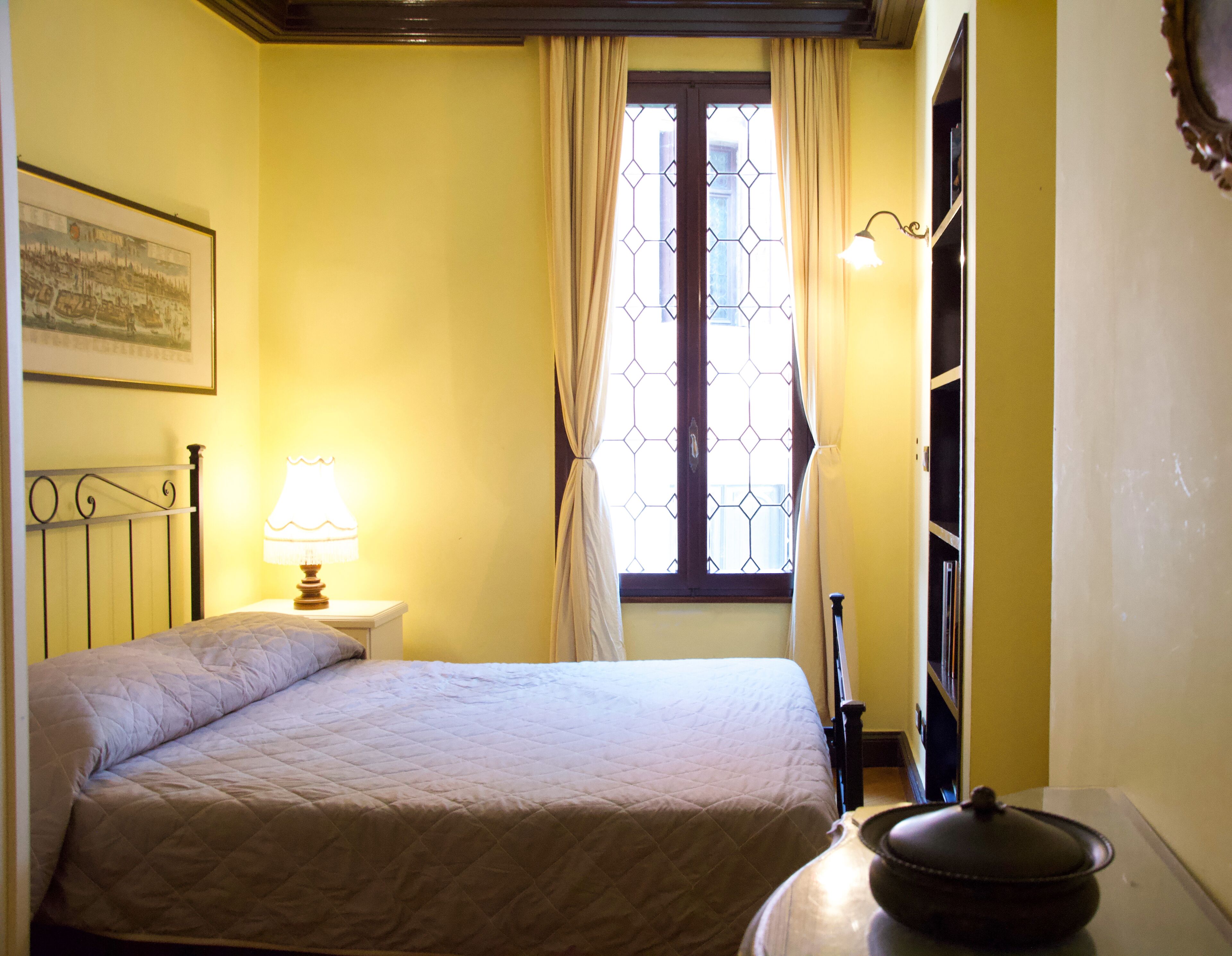 Habitació amb un llit, una tauleta de nit amb una làmpada, una finestra amb cortines i una estanteria penjada a la paret.