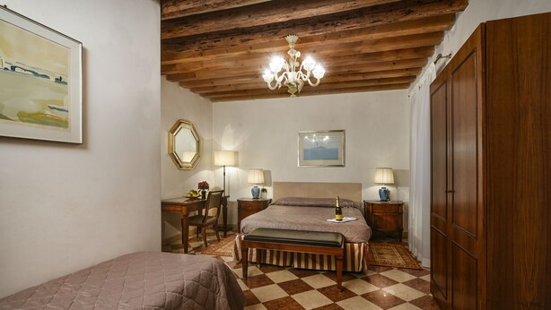 A bedroom with a checkered floor, wooden ceiling beams, a chandelier, two beds, bedside tables with lamps, a desk with a chair, and a large wardrobe. - Anzolo Palace (Venice)