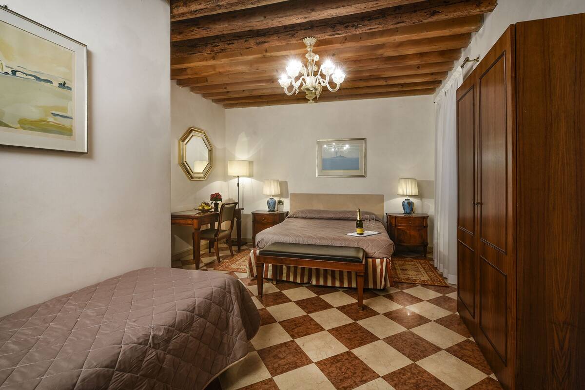 Un dormitorio con suelo a cuadros, vigas de madera en el techo, un candelabro, dos camas, mesitas de noche con lámparas, un escritorio con silla y un armario grande.