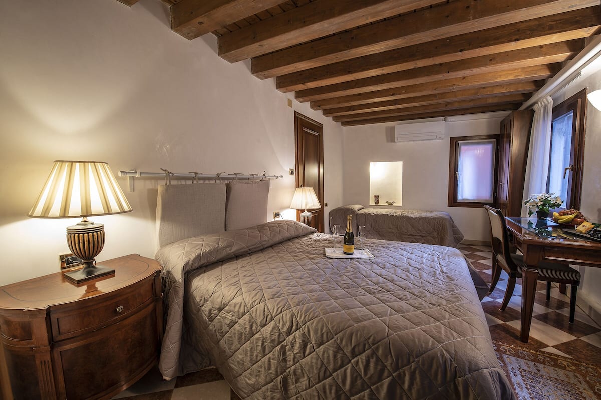 Un dormitorio con dos camas, vigas de madera en el techo, una mesita de noche con una lámpara y un pequeño escritorio con una planta y fruta fresca.