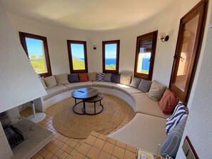 Ampla sala de estar com sofá curvo, mesa de centro redonda e vista para o mar através de grandes janelas.