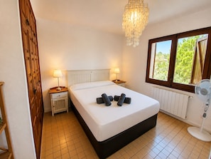 A hotel room with a bed, two bedside tables with lamps, a radiator, a window with a view of greenery, and a standing fan.