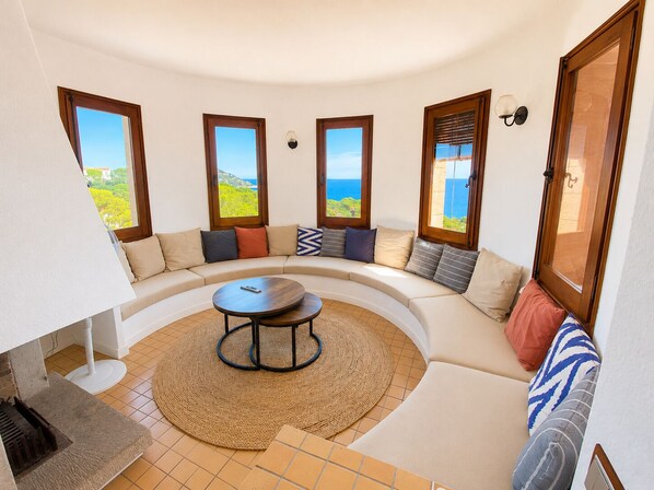 Sala de estar circular com um sofá, uma mesa de centro e vista para o mar através de grandes janelas.