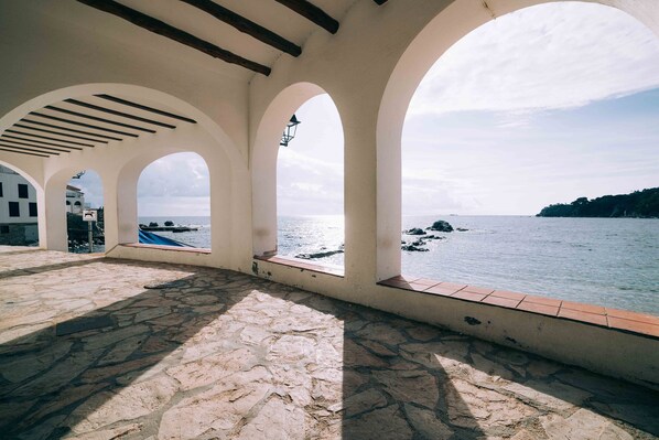 Um terraço de pedra com janelas em arco, com vista para um corpo d'água e uma linha de costa distante.