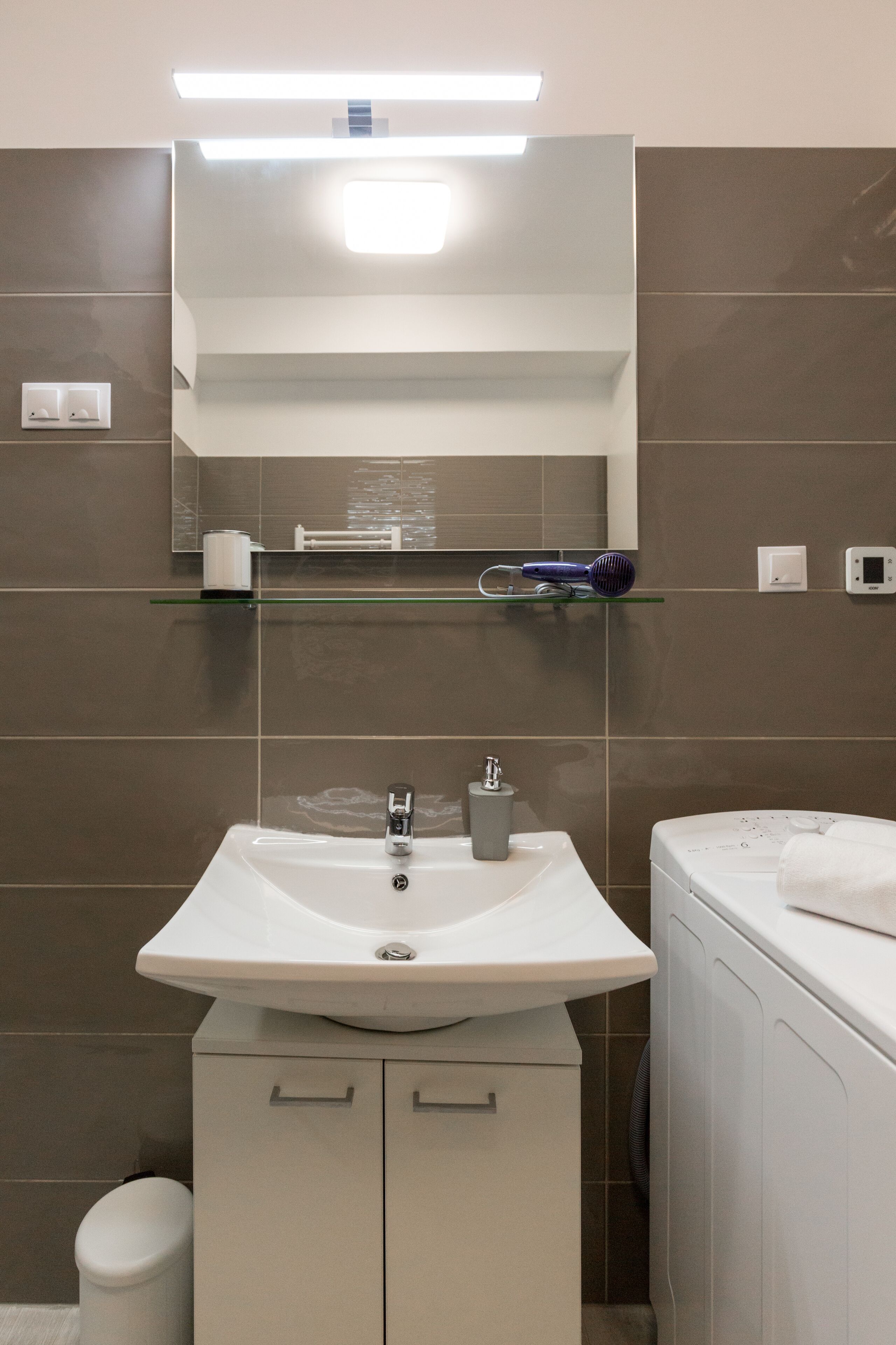 Une salle de bain moderne avec un lavabo blanc, un miroir et une étagère en verre au-dessus du miroir.