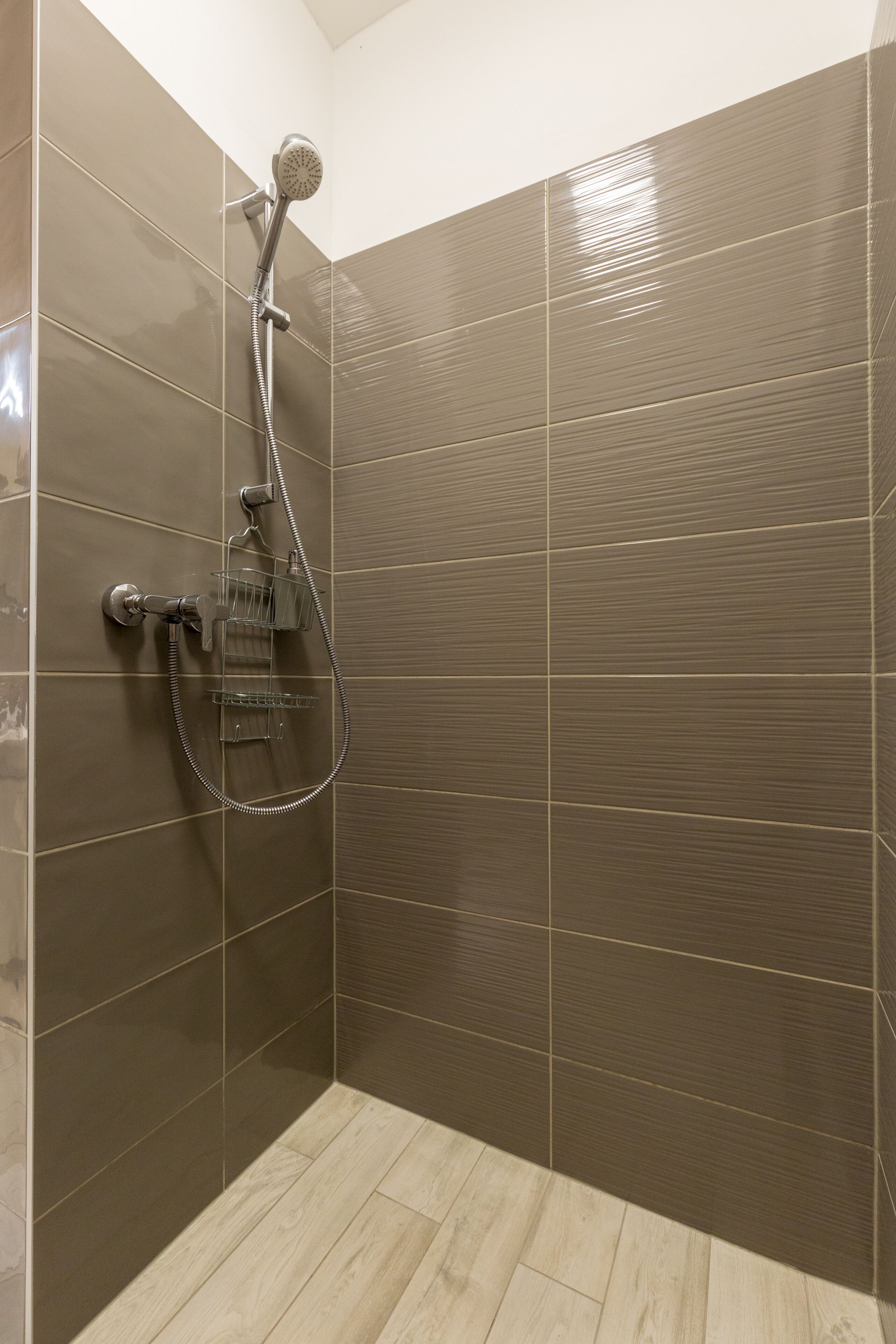 A shower with a handheld showerhead and a wall-mounted faucet, tiled walls, and a wooden floor.