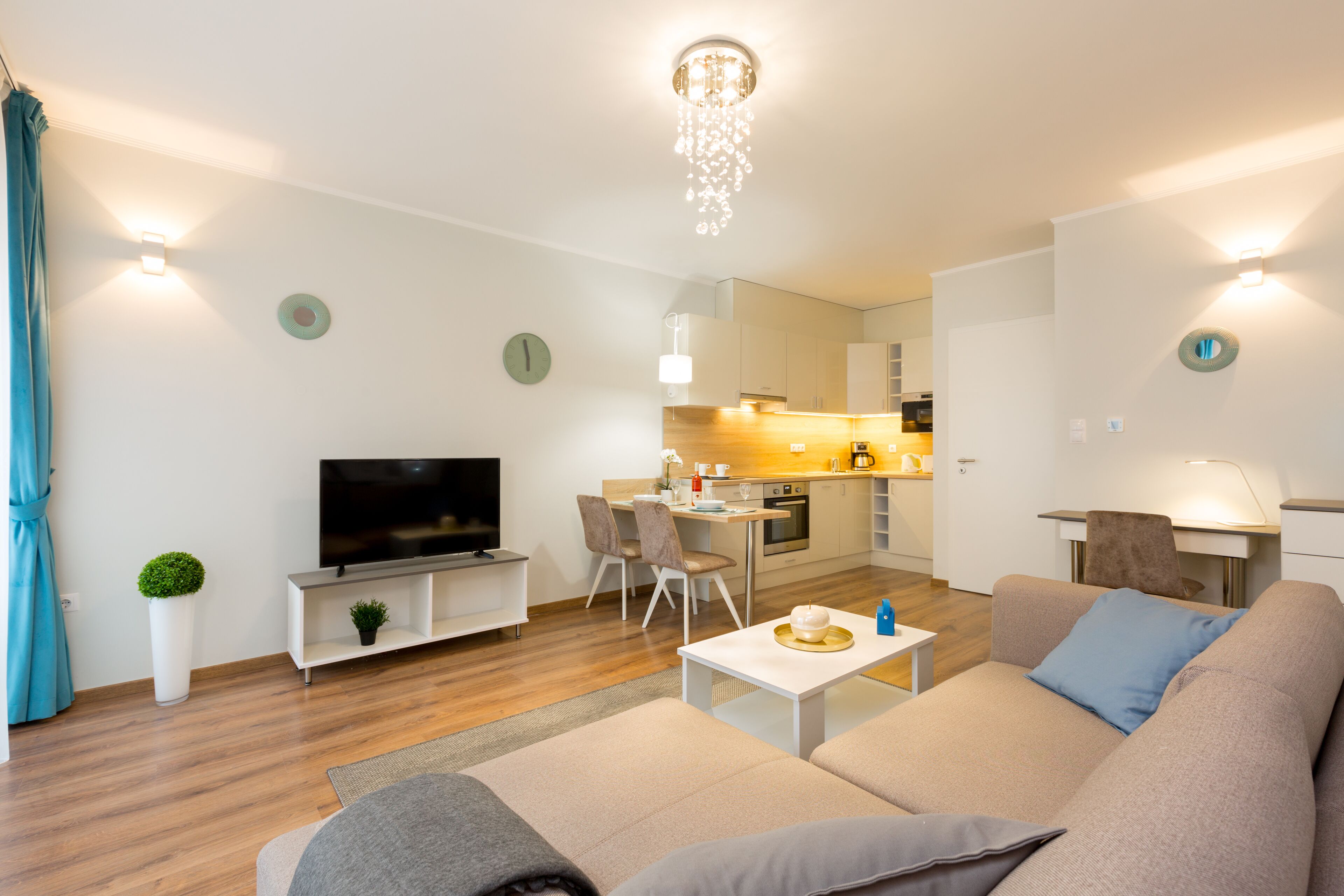 A modern living room with a sofa, a coffee table, and a TV. There is a kitchen in the background with a dining area.
