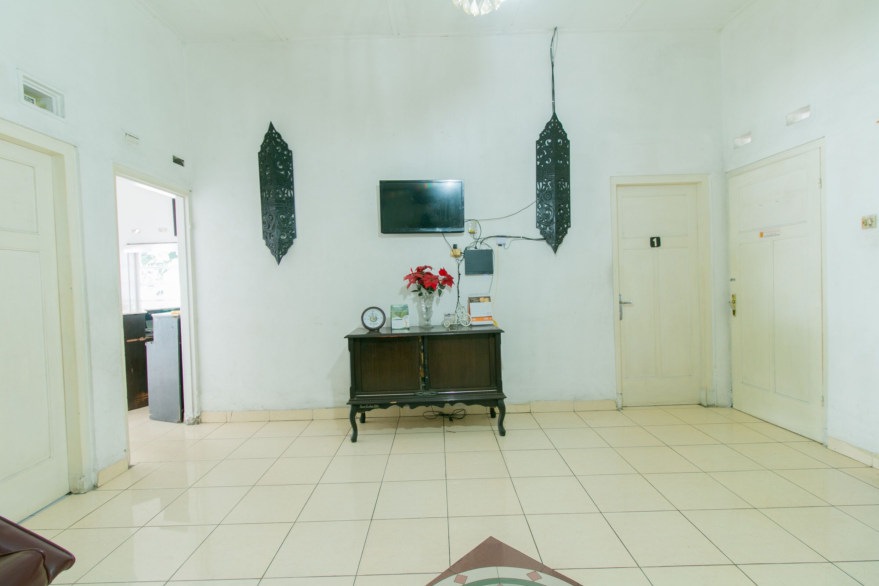 Una habitación amplia con suelo de baldosas, una consola de madera oscura con adornos, un televisor de pantalla plana en la pared y una puerta con un número.