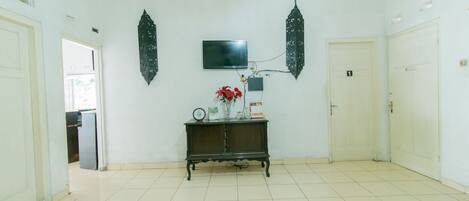 Una habitación amplia con suelo de baldosas, una consola de madera oscura con adornos, un televisor de pantalla plana en la pared y una puerta con un número.