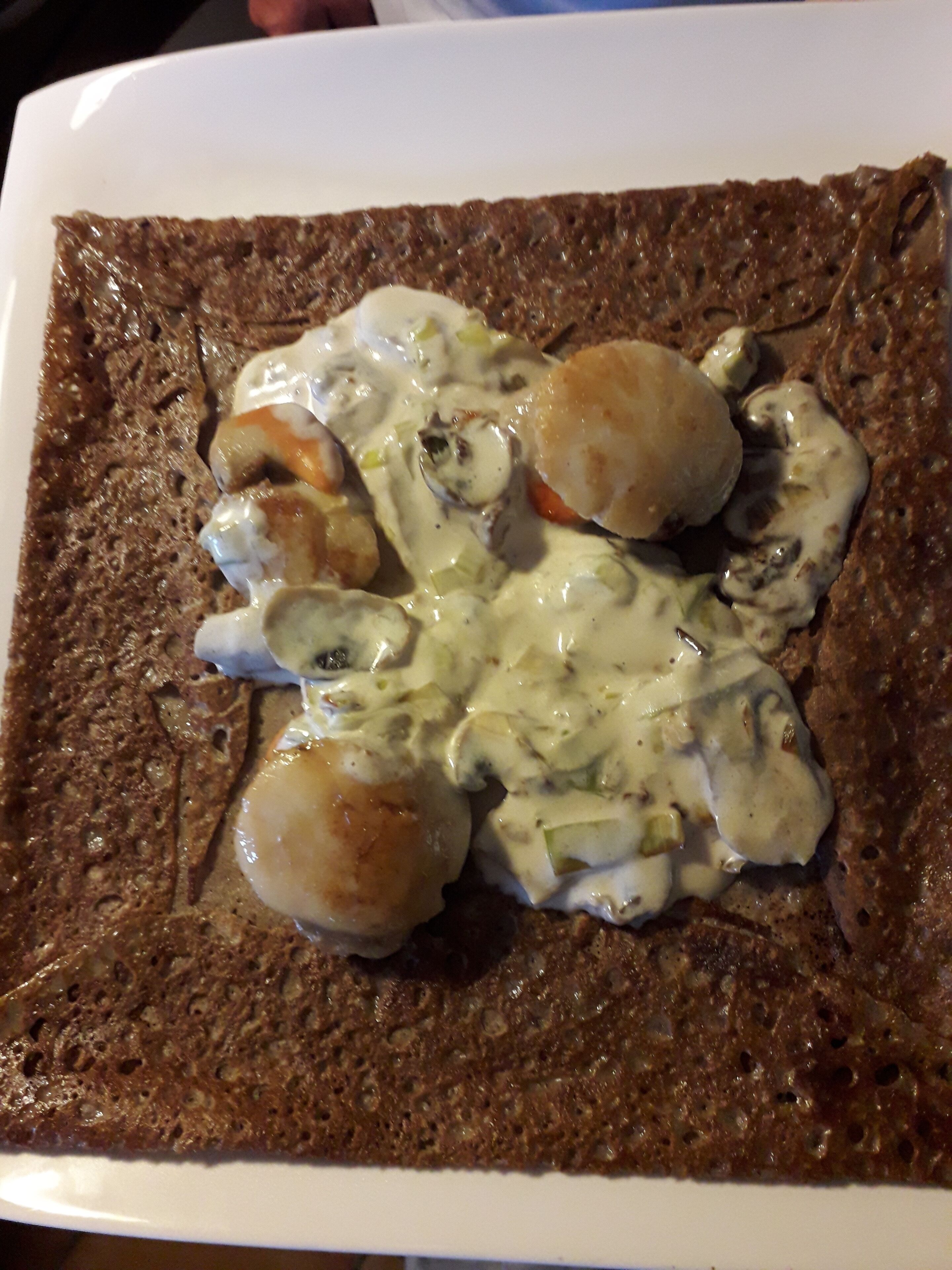 En firkantet tallerken med en mørkebrun, tekstureret spise, muligvis en fladbrød, toppet med en cremet sauce og flere runde, lysebrune objekter, sandsynligvis muslinger eller rejer.