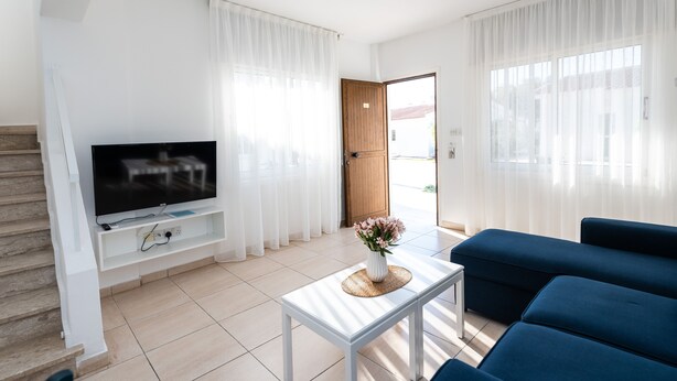 A modern living room with a flat-screen TV on a white stand, a blue sofa, a coffee table with a vase of flowers, and a staircase to the left. - Fig Tree Z Holiday Villas DPS02- Two Bed Townhouse (Protaras)