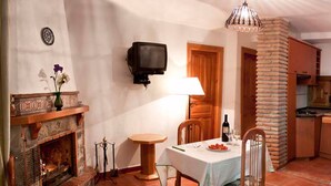 Un salon chaleureux avec une cheminée, un téléviseur fixé au mur, une table à manger avec des chaises et un coin cuisine agrémenté d’un pilier en briques.