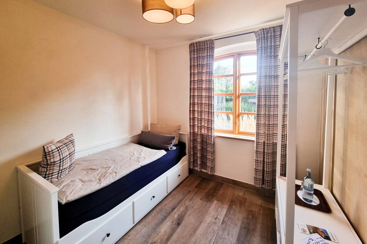 Une chambre à coucher avec un lit, une fenêtre avec des rideaux, un radiateur et une petite table sur laquelle se trouve une bouteille.