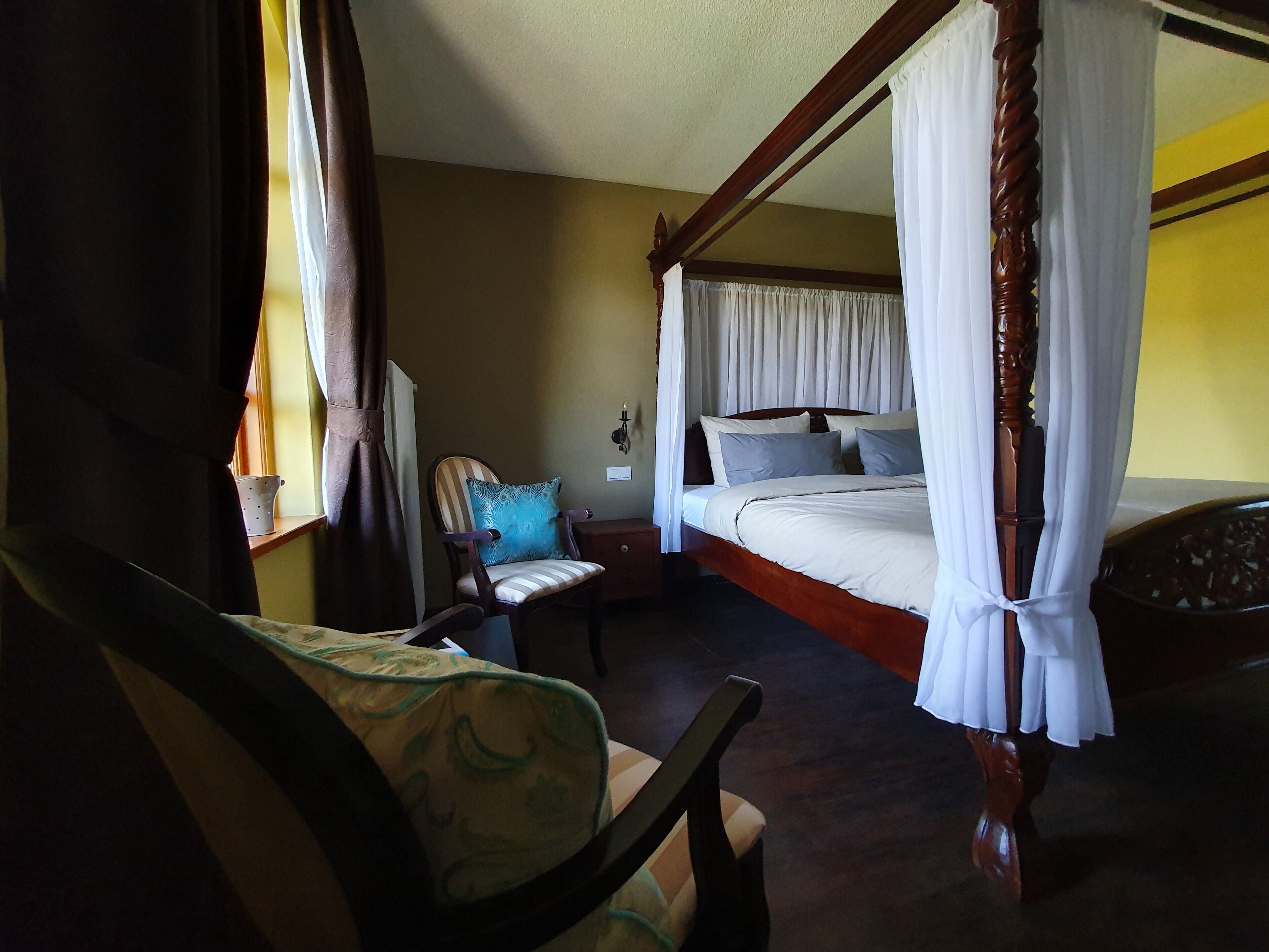 Uma cama com dossel, cortinas brancas, uma mesinha de cabeceira de madeira, uma cadeira com almofada estampada e uma janela com cortinas.