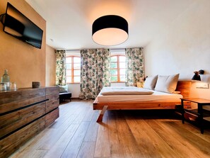 Un dormitorio con una cama de madera, una mesita de noche, una silla, un escritorio, un televisor montado en la pared y una ventana con cortinas.