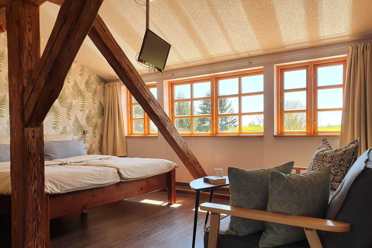 Une chambre avec un lit en bois, une petite table, un canapé agrémenté de coussins à motifs et de grandes fenêtres donnant sur un paysage verdoyant.