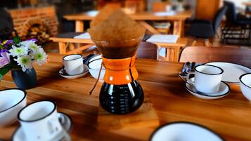 Una cafetera sobre una mesa de madera con tazas y platillos, un florero con flores, y una pared de ladrillo al fondo.