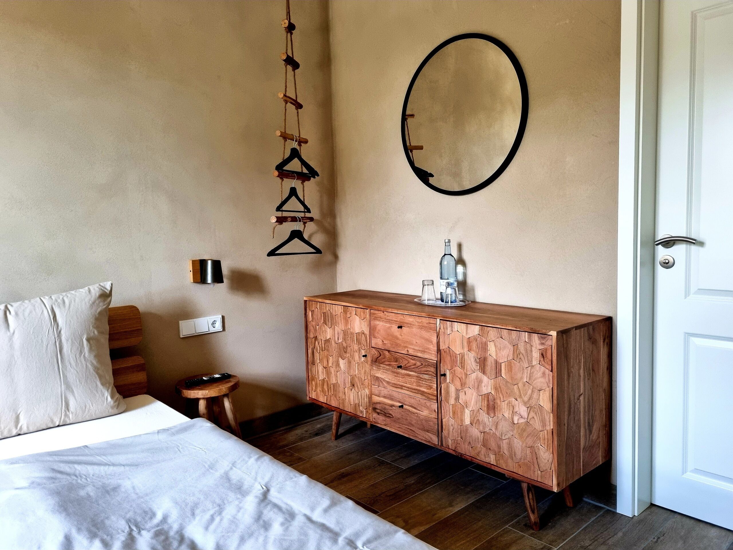 Un dormitorio con un aparador de madera, una cama con ropa blanca, una lámpara colgante y un espejo redondo en la pared.