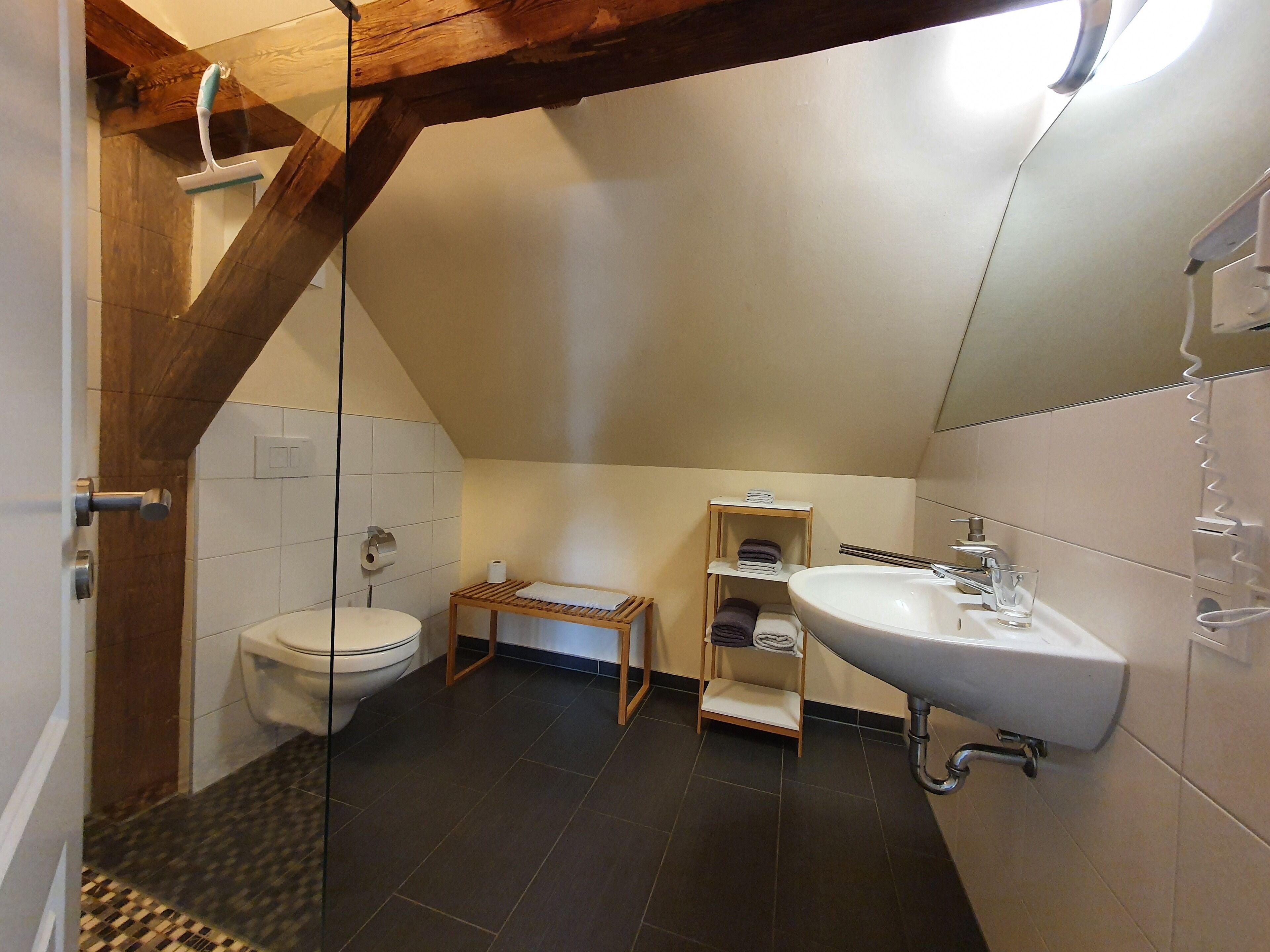 Et lille, moderne badeværelse med toilet, vask og brusebad. Badeværelset har mørke fliser, træbjælker og en lille hylde med håndklæder.