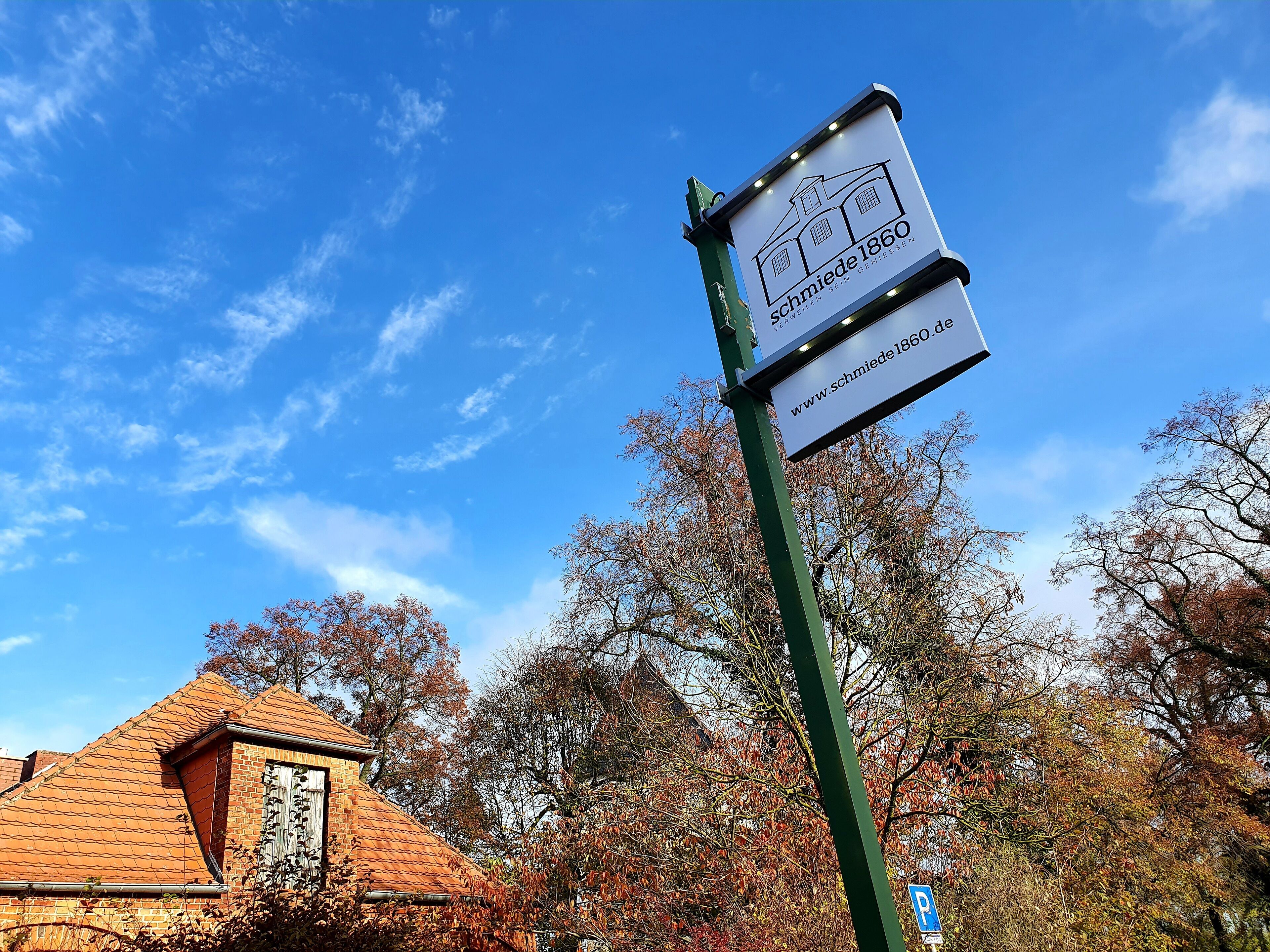 A sign for 'Schmiede 1860' with a website URL, set against a clear blue sky and autumn trees.